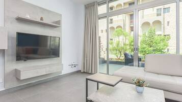 A modern living room with a flat-screen TV, a sofa, a coffee table, and a balcony with a view of a building and greenery.