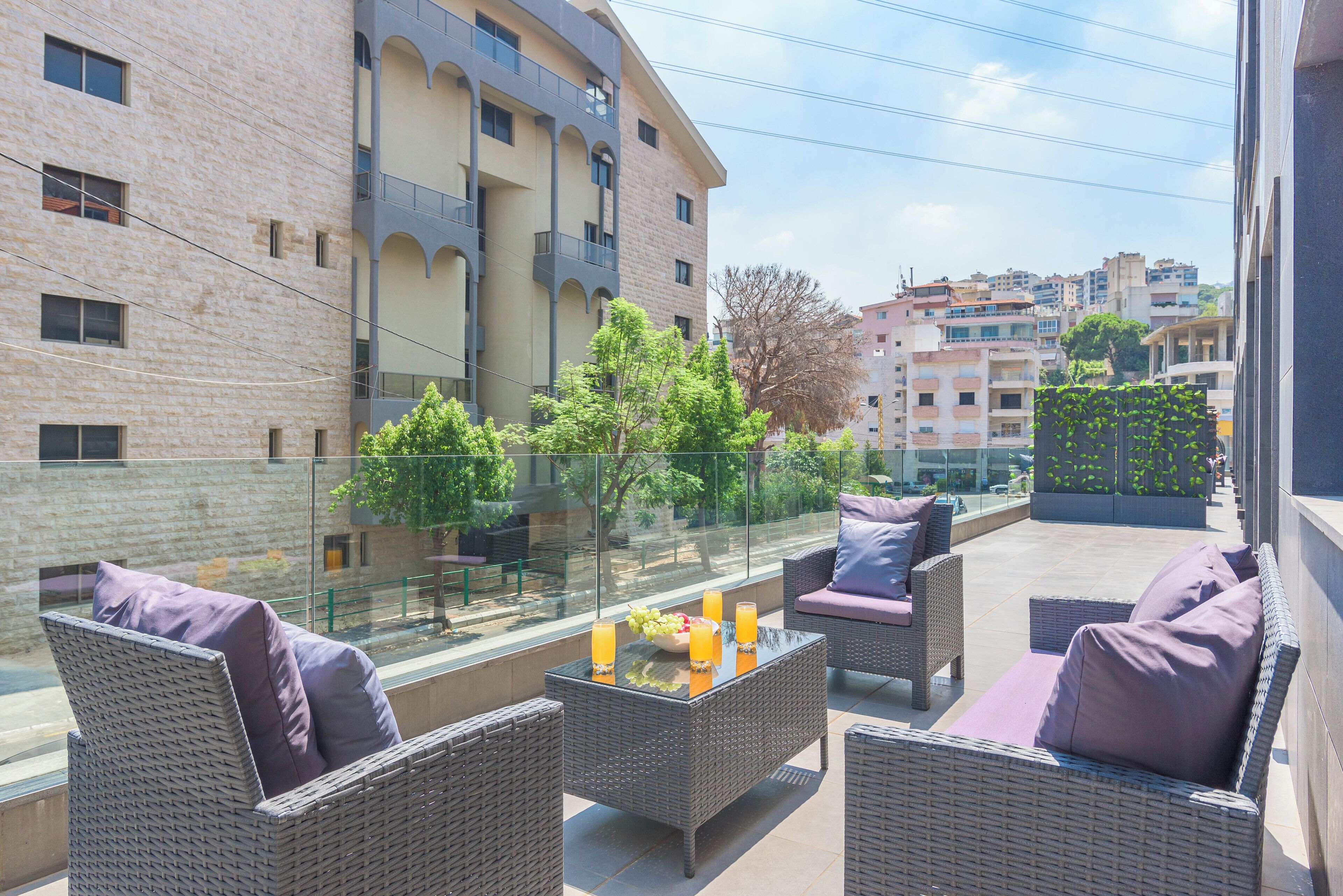 A balcony with wicker furniture, a glass table with fruit and drinks, and a view of surrounding buildings.
