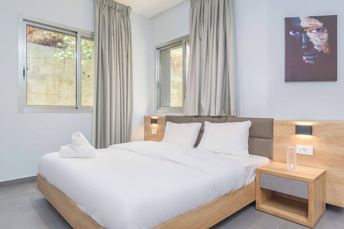 A modern bedroom with a large bed, two windows with curtains, a headboard with integrated lighting, and a bedside table with a glass container.