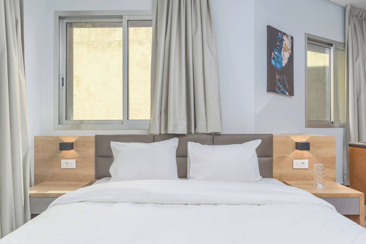 A modern hotel room with a large bed, two bedside tables, a nightstand, a window with curtains, and a wall-mounted artwork.