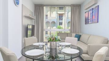 Una sala de estar moderna con una mesa de comedor de cristal preparada para cuatro personas, un sofá color beige y un ventanal con cortinas.