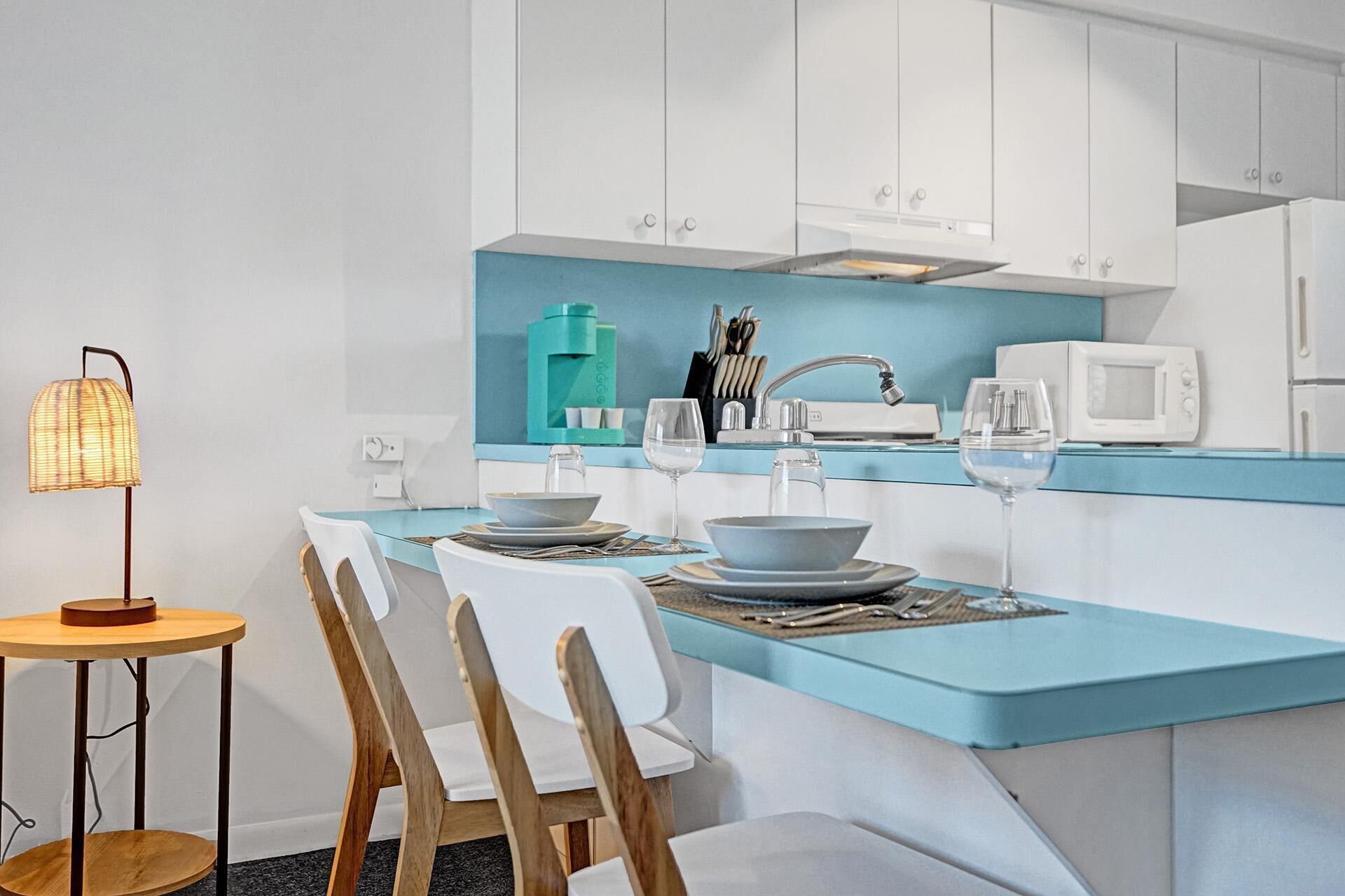 Une cuisine moderne avec des meubles blancs, un plan de travail de couleur turquoise et un coin repas comprenant des chaises blanches et une petite table ronde.