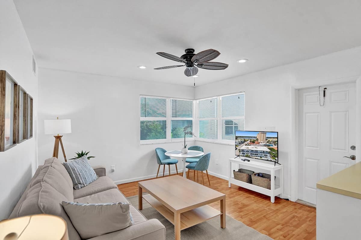 A modern living room with a sofa, a coffee table, and a TV. There are windows with blinds, a ceiling fan, and a door.