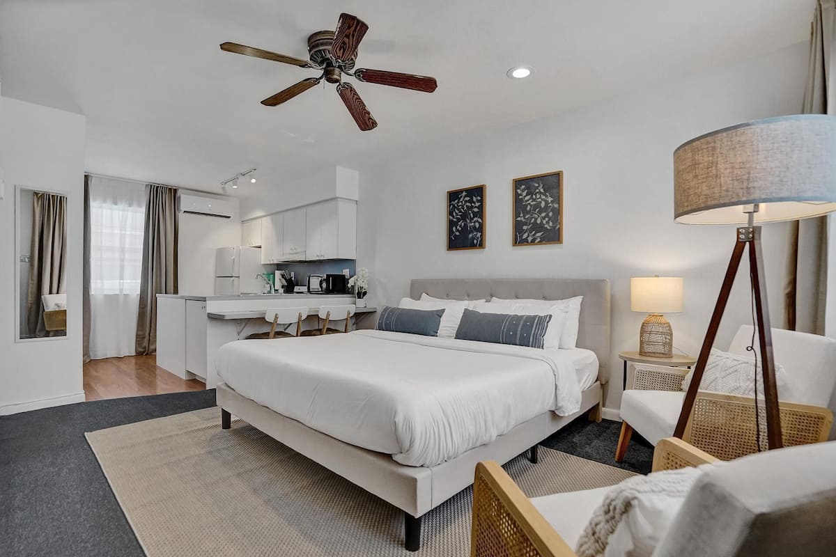 A modern hotel room with a large bed, a desk, a kitchenette, and a ceiling fan.