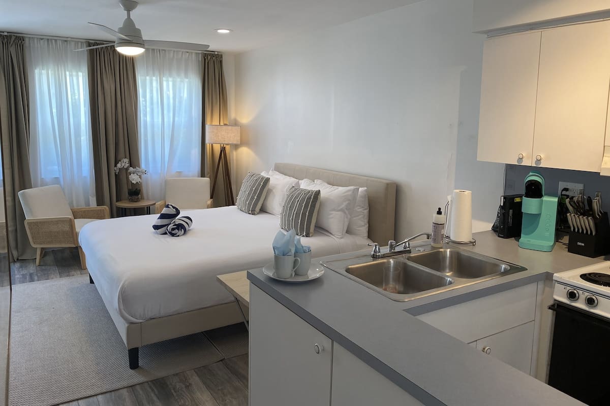 A hotel room with a bed, bedside table, lamp, chair, and kitchenette.