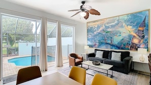 Een moderne woonkamer met een groot raam, een plafondventilator, een schilderij van een stadsgezicht, een zwembad en een eethoek.