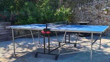 Ein Outdoor-Tischtennis-Tisch auf einer Betonfläche, im Hintergrund eine Steinmauer und Bäume.
