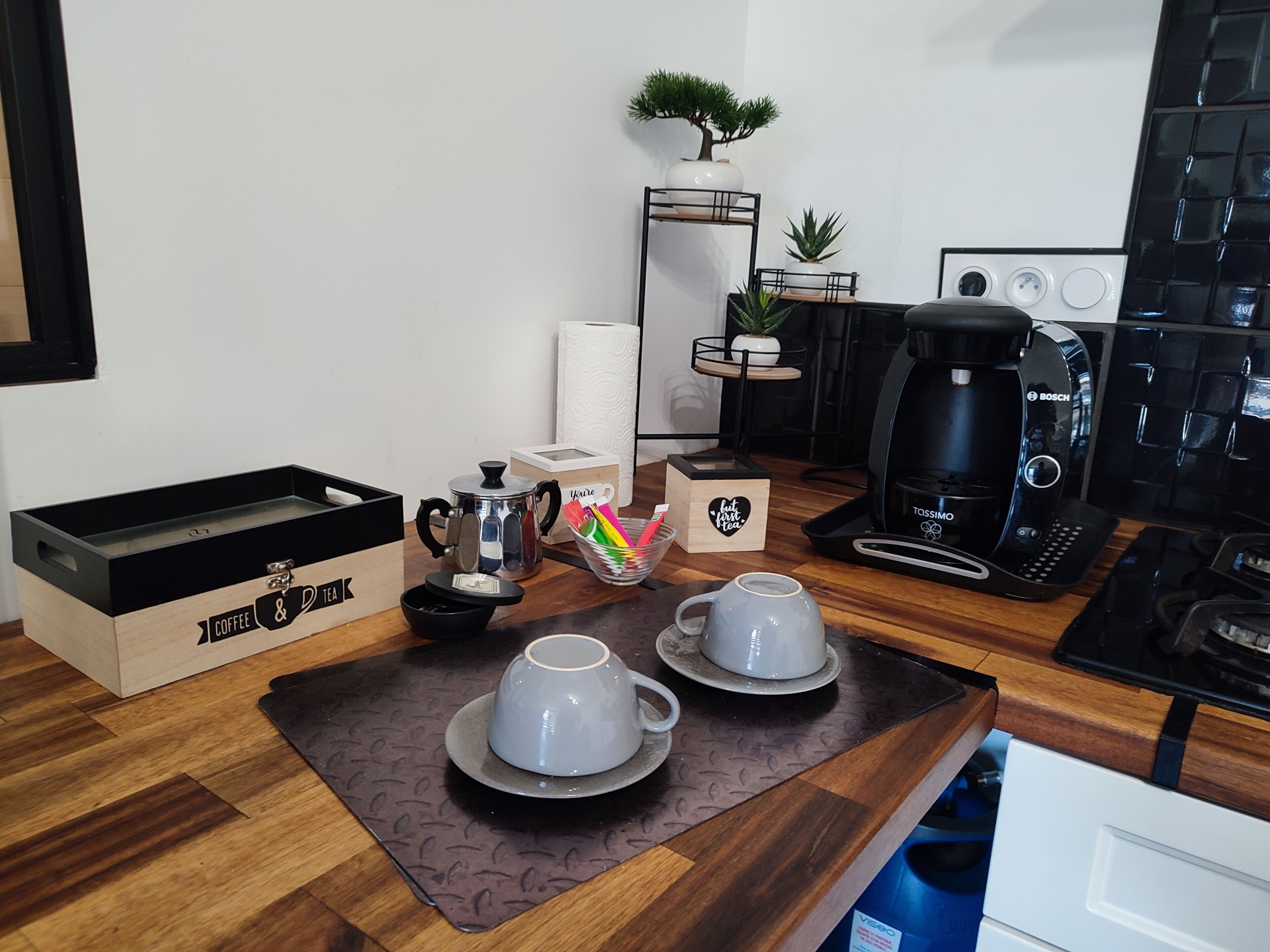 A compact kitchen with a coffee maker, two cups, a tea box, and a small plant.