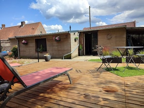 A wooden deck with a red chair, a table, and potted plants. There is a small building with a tiled roof and a door.