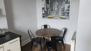 A dining area with a round wooden table, four black chairs, a white cabinet with drawers, and a cityscape painting on the wall. - Snug - Abhainn Apartment (Helensburgh)