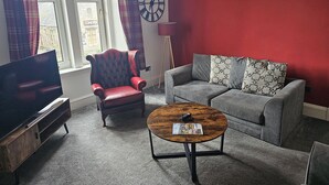 A living room with a red wall, a grey sofa, a red armchair, a wooden coffee table, and a television. - Snug - Abhainn Apartment (Helensburgh)