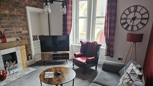 A living room with a fireplace, a television, a red leather armchair, a grey sofa, and a large wall clock. - Snug - Abhainn Apartment (Helensburgh)