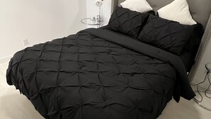 Een slaapkamer met een groot bed, een grijs gestoffeerd hoofdeinde en een zwart gewatteerde sprei.