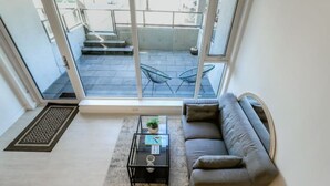 Een moderne woonkamer met een grijze bank, een glazen salontafel en uitzicht op een balkon met buitenmeubilair.