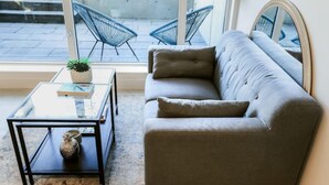 Een moderne woonkamer met een grijze bank, een glazen salontafel met een boek en een plant, en twee blauwe stoelen voor een groot raam.