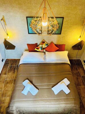 A bed with a geometric-patterned pillow, a hanging geometric light fixture, and a framed artwork on the wall.