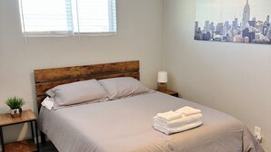 Une chambre à coucher bien rangée, avec un lit, des tables de chevet, un ventilateur de plafond, une fenêtre avec des stores et un tableau représentant un paysage urbain accroché au mur.