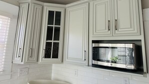 Una cocina con armarios blancos, un microondas de acero inoxidable y una ventana con persianas.