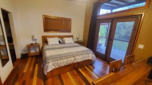 Un dormitorio con cama de madera, una mesita de noche con lámpara, una ventana con persianas y vista a una terraza.