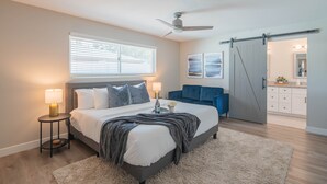 A bedroom with a bed, bedside tables, a ceiling fan, a large window with blinds, a sliding door leading to a bathroom, and a light-colored rug. - Centrally Located Hike Dine & Shop w/ Pool Table, Heated Pool, Office, Firepit (Phoenix)