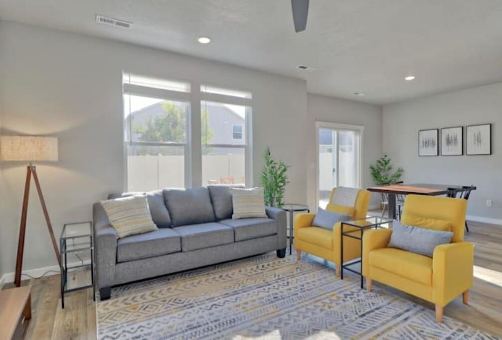 A modern living room with a grey sofa, a yellow armchair, and a patterned rug. There is a dining table with chairs, a lamp, and a plant.