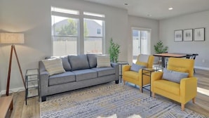 Una sala moderna con un sofá gris, un sillón amarillo y una alfombra estampada. Se observa una mesa de comedor con sillas, una lámpara y una planta.