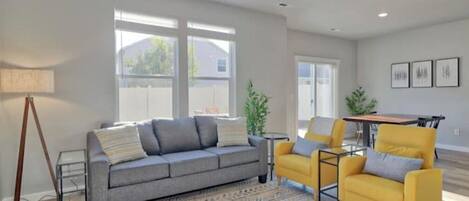A modern living room with a grey sofa, a yellow armchair, and a patterned rug. There is a dining table with chairs, a lamp, and a plant.