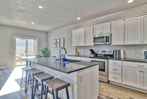 A modern kitchen with white cabinets, a central island with stools, stainless steel appliances, and a herringbone tile backsplash.