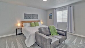 Un dormitorio con cama, un sofá gris, una mesita de noche y una ventana con cortinas.