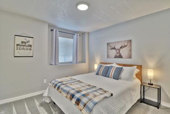 A bedroom with a bed, a nightstand, a window with blinds, and two wall decorations.