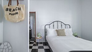 Un dormitorio con una cama, una bolsa de playa colgante, una mesita auxiliar pequeña y una silla.
