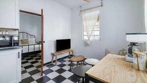 Una cocina compacta con piso a cuadros blanco y negro, una mesa de comedor de madera, un televisor pequeño sobre un soporte y un ventilador de techo.