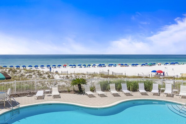 A pool area with lounge chairs and umbrellas, a beach with umbrellas and people, and a clear sky.