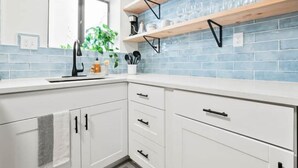 Una cucina con mobili bianchi, una finestra, una pianta e uno schienale piastrellato blu.