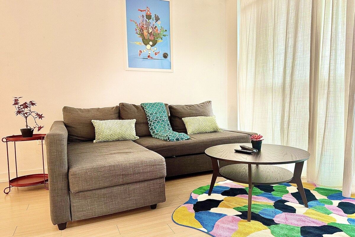 Ein Wohnzimmer mit einer Couch, einem Couchtisch, einem farbenfrohen Teppich, einem kleinen Beistelltisch mit Pflanze und einem Gemälde an der Wand.