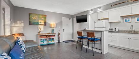 Een moderne keuken met witte kasten, een ingebouwde magnetron en een flatscreen televisie. Er is een eethoek met twee rieten stoelen en een kleine tafel.