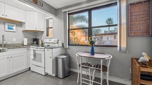 Cocina compacta con alacenas blancas, estufa y un cubo de basura. Hay una mesa pequeña con una silla y un jarrón de flores. Una ventana con persianas ofrece vistas a palmeras y edificios.