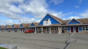 Un motel con una hilera de habitaciones, un coche rojo estacionado al frente y un cielo azul despejado.