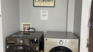 Dos lavadoras de carga frontal en un cuarto de lavado con un letrero que dice "LAUNDRY WASH, DRY & FOLD" y un aviso de servicio de lavandería a $10 en efectivo.
