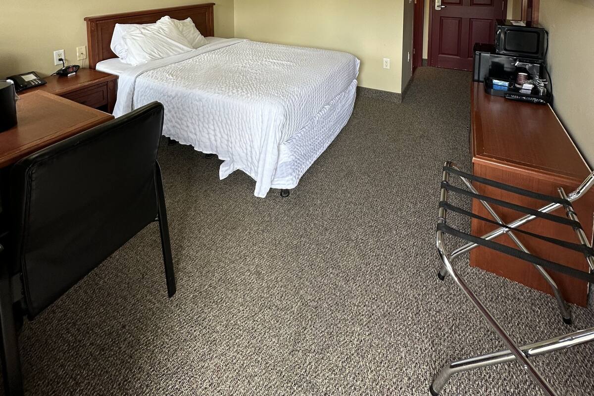 A hotel room with a bed, a desk, a chair, a ceiling fan, a door, and a small kitchenette.
