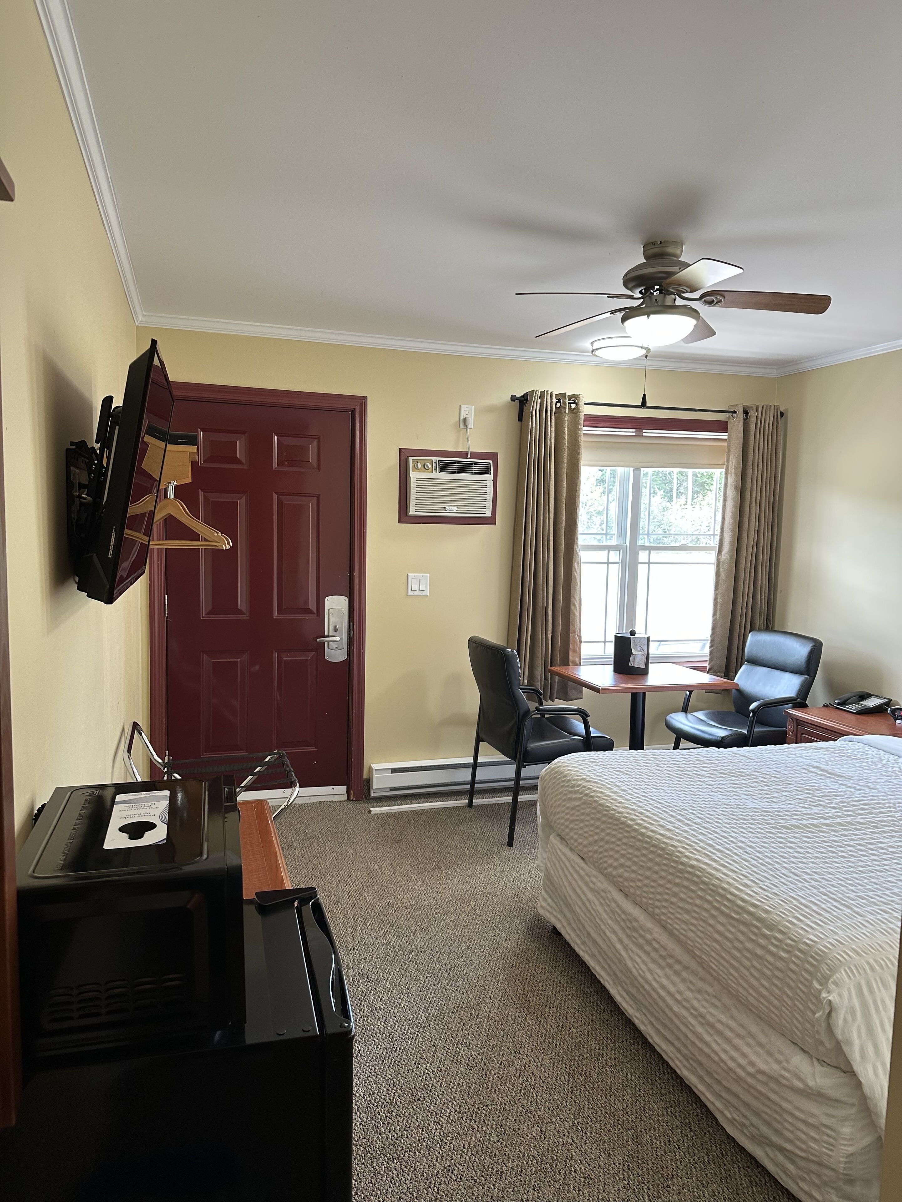 Quarto de hotel com cama, escrivaninha, cadeiras, televisão e ventoinha no teto.