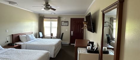 A hotel room with two beds, a desk, a trash bin, a ceiling fan, and a large mirror.