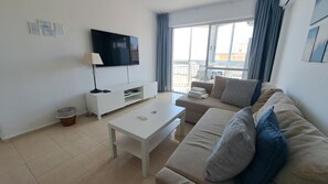 A living room with a TV, white furniture, a balcony with a view, and a trash bin.