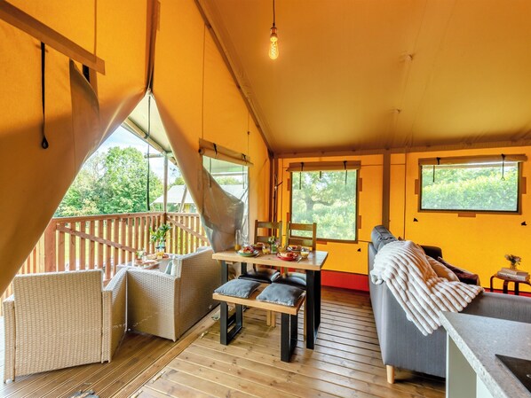 A cozy, well-lit room with a wooden floor, a dining table set with food and drinks, a sofa, and a window overlooking greenery.