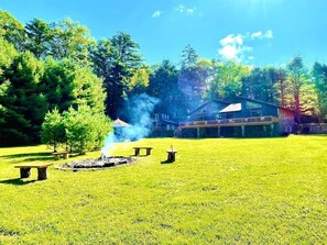 Un espacio al aire libre con césped, un hogar de piedra, bancos y una casa al fondo.