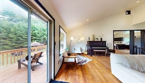 Amplia sala de estar con un gran ventanal, un piano, un sofá y una terraza de madera.