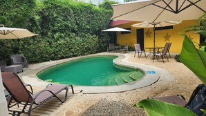 A swimming pool with a curved edge, surrounded by lounge chairs and umbrellas, with a yellow wall and greenery in the background.