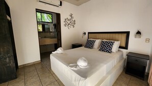 A hotel room with a bed, a nightstand, a bedside table, a wall-mounted lamp, a decorative wall piece, a bathroom with a sink and mirror, and a window with a view of greenery.