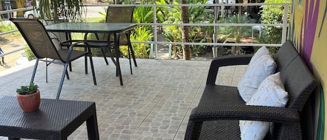 An outdoor patio with a wicker sofa, a table, and chairs, surrounded by greenery and a view of the street.