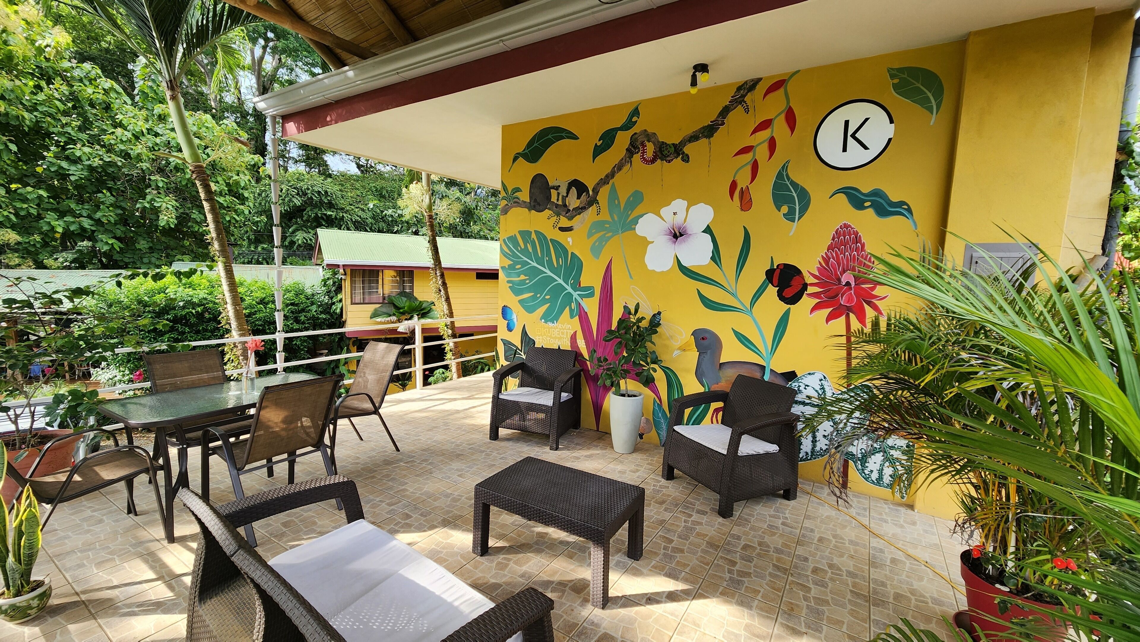 An outdoor patio with wicker furniture, a mural with tropical plants, and a yellow wall with a 'K' symbol.